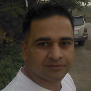A man with short dark hair, wearing a white shirt, stands outdoors near a road with lush greenery and a parked car in the background. Sourcing, Procurement, 3rd Party Risk