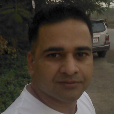 A man with short dark hair, wearing a white shirt, stands outdoors near a road with lush greenery and a parked car in the background. Sourcing, Procurement, 3rd Party Risk
