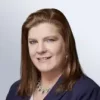 A woman with straight, shoulder-length brown hair is wearing a navy blazer and a necklace, smiling confidently at the camera against a light gray background. Sourcing, Procurement, 3rd Party Risk