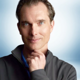 A man with light skin and blue eyes, wearing a dark zip-up jacket over a blue shirt, poses thoughtfully with his hand on his chin against a serene light blue background. Sourcing, Procurement, 3rd Party Risk