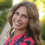 A woman with wavy light brown hair, wearing a pink and navy floral top, smiles while standing outdoors on a sunny day surrounded by lush greenery. Sourcing, Procurement, 3rd Party Risk