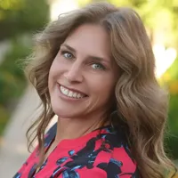 A woman with wavy light brown hair, wearing a pink and navy floral top, smiles while standing outdoors on a sunny day surrounded by lush greenery. Sourcing, Procurement, 3rd Party Risk