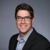 A man with short brown hair and glasses, wearing a dark blazer over a light blue collared shirt, confidently smiles at the camera against a plain gray background. Sourcing, Procurement, 3rd Party Risk
