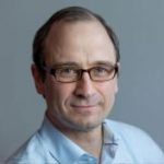 A man with short brown hair and glasses is wearing a light blue collared shirt, looking at the camera with a slight smile against a plain grey backdrop. Sourcing, Procurement, 3rd Party Risk