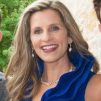 A woman with long, wavy blonde hair is smiling. She wears a royal blue dress with ruffled details, drop earrings, and a necklace. The lush greenery and a stone wall create an elegant backdrop. Sourcing, Procurement, 3rd Party Risk