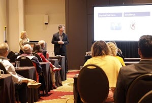 A group of people seated in a conference room listen to two presenters near a large, illuminated screen showing a digital presentation. Sourcing, Procurement, 3rd Party Risk