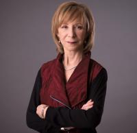 A woman with light brown hair stands against a gray background, wearing a maroon vest over a black top, her arms crossed and a neutral expression, exuding subtle confidence. Sourcing, Procurement, 3rd Party Risk