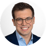 A man with short brown hair, wearing round eyeglasses, a light blue shirt, and a dark blazer, smiles confidently at the camera against a plain light background. Sourcing, Procurement, 3rd Party Risk