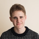 A young person with short light brown hair, wearing a dark sweater with a white patterned design, is smiling softly in front of a plain light-colored background, exuding warmth and approachability. Sourcing, Procurement, 3rd Party Risk