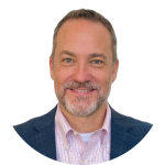 A middle-aged man with short brown hair, a trimmed gray beard, and blue eyes is smiling. He wears a blue blazer over a pink checkered shirt and white undershirt. The background is plain white, highlighting his approachable style. Sourcing, Procurement, 3rd Party Risk