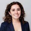 A woman with shoulder-length curly brown hair, wearing a navy blazer and blue earrings, smiles at the camera in front of a minimalist light gray background. Sourcing, Procurement, 3rd Party Risk