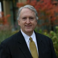 A middle-aged man with gray hair, wearing a dark suit, white shirt, and yellow patterned tie, stands outdoors amid blurred autumn foliage and vibrant leaves in the background. Sourcing, Procurement, 3rd Party Risk