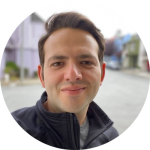 A person with short brown hair and fair skin smiles slightly while standing outdoors on a street, wearing a dark zip-up jacket. The softly blurred background features city buildings and trees. Sourcing, Procurement, 3rd Party Risk