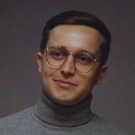 A person with short brown hair, wearing round glasses and a stylish gray turtleneck sweater, smiles softly against a plain, dark background. Sourcing, Procurement, 3rd Party Risk