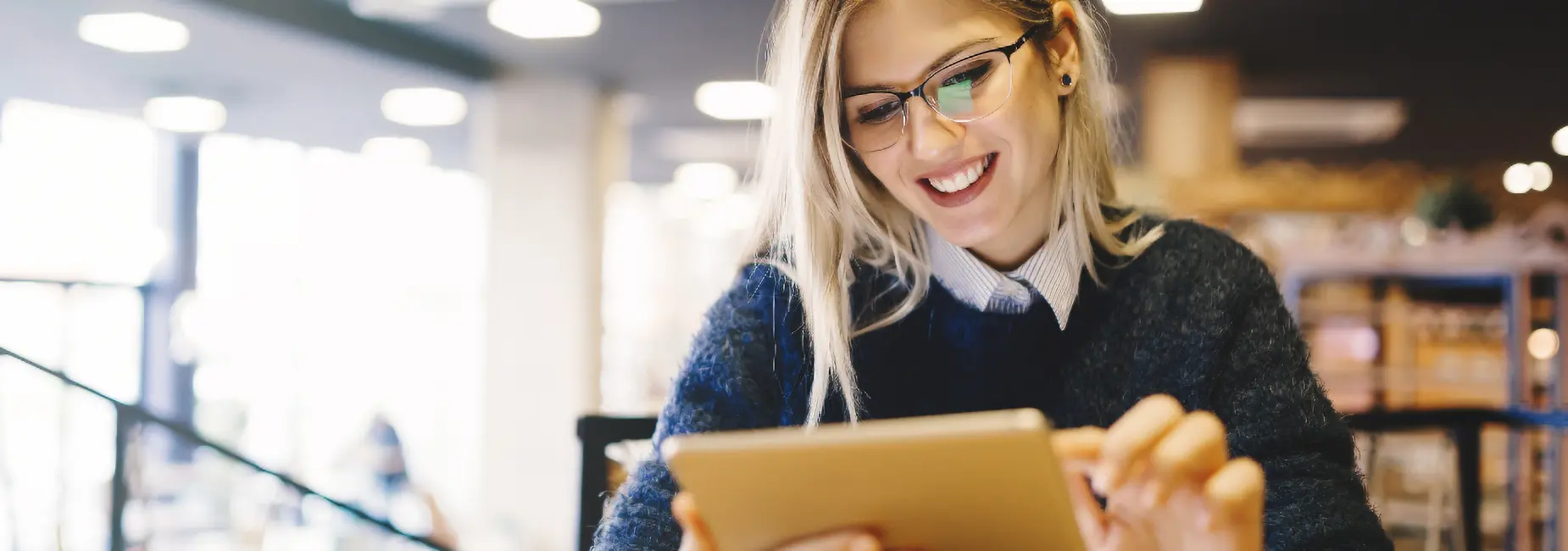 A smiling woman wearing glasses and a dark sweater sits indoors at a table, using a tablet device. The softly blurred background suggests a cozy, well-lit café or modern workspace. Sourcing, Procurement, 3rd Party Risk