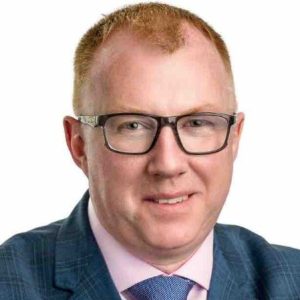 A man with short red hair and glasses wears a blue plaid suit, pink shirt, and blue tie, smiling confidently at the camera against a white background. Sourcing, Procurement, 3rd Party Risk