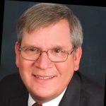 Smiling older man with short gray hair, wearing glasses, a dark suit, white shirt, and patterned tie, confidently posing in front of a dark background. Sourcing, Procurement, 3rd Party Risk