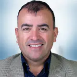 A man with short dark hair and a receding hairline is smiling. He is wearing a checked blazer over a dark shirt. The softly blurred background features a light blue and white gradient, adding a professional touch to the portrait. Sourcing, Procurement, 3rd Party Risk