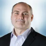 A man with short hair, wearing a dark suit jacket and light-colored dress shirt, poses confidently in front of a softly blurred blue and white background, smiling slightly. Sourcing, Procurement, 3rd Party Risk