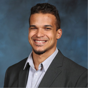 A man with short curly hair and a trimmed beard smiles while wearing a dark gray suit jacket over a light gray collared shirt, set against a blue and gray blurred background for a professional look. Sourcing, Procurement, 3rd Party Risk