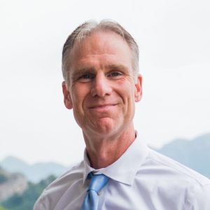 A middle-aged man with short gray hair smiles at the camera, wearing a white dress shirt and a light blue tie. The blurred background features scenic mountains and a cloudy sky. Sourcing, Procurement, 3rd Party Risk
