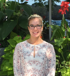 A woman with blond hair in a bun, wearing glasses and a light-colored floral blouse, smiles warmly while standing in front of lush green plants and vibrant red flowers outdoors. Sourcing, Procurement, 3rd Party Risk