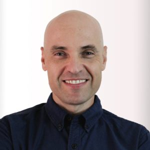 A smiling bald man in a dark button-up shirt stands against a minimalist, light-colored background. Sourcing, Procurement, 3rd Party Risk