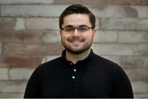 A young man with short dark hair, glasses, and a trimmed beard smiles at the camera. He is wearing a black collared shirt and stands in front of a modern light gray brick wall background. Sourcing, Procurement, 3rd Party Risk