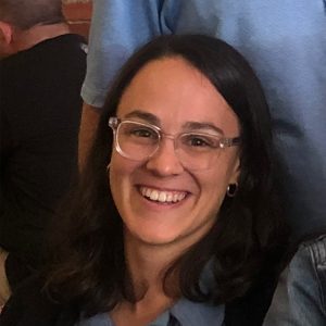 A woman with shoulder-length dark hair, wearing clear glasses and a blue top, is smiling indoors. Another person in a light blue shirt is partially visible behind her. The scene feels friendly and inviting. Sourcing, Procurement, 3rd Party Risk