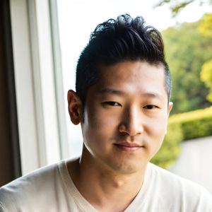 A young man with short, styled black hair and a slight smile is indoors near a window, wearing a light-colored V-neck shirt. Sunlight filters through the window, revealing green trees and shrubbery outside. Sourcing, Procurement, 3rd Party Risk