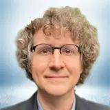 A person with curly light brown hair, glasses, and a slight smile, wearing a suit jacket, is pictured against a blurred blue and white background, exuding a professional and approachable demeanor. Sourcing, Procurement, 3rd Party Risk