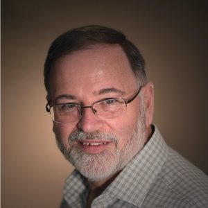A middle-aged man with short brown hair, a beard, and glasses smiles at the camera. He is wearing a light-colored checkered shirt and is posed against a soft brown gradient background. Sourcing, Procurement, 3rd Party Risk