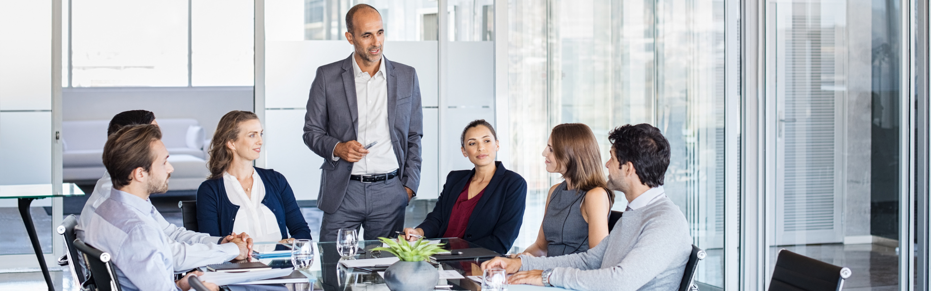 Six business professionals sit around a conference table in a modern office, attentively listening as a suited manager presents new ideas. Sourcing, Procurement, 3rd Party Risk