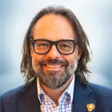 A smiling man with long brown hair, a beard, and glasses, wearing a dark suit jacket over a checkered shirt, stands against a softly blurred light blue background. Sourcing, Procurement, 3rd Party Risk