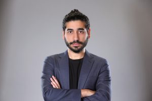 A man with dark hair and a beard, wearing a dark blazer and black shirt, stands with arms crossed against a plain gray background, exuding confidence as he looks directly at the camera. Sourcing, Procurement, 3rd Party Risk