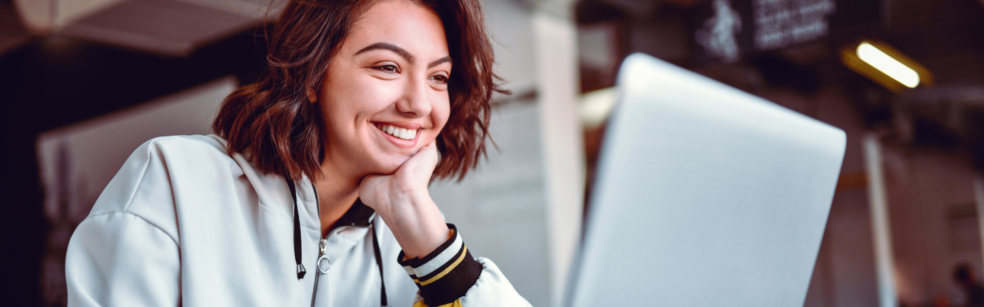A young woman with shoulder-length brown hair smiles while looking at a laptop screen in a bright indoor setting, appearing engaged and happy as she explores new ideas. Sourcing, Procurement, 3rd Party Risk