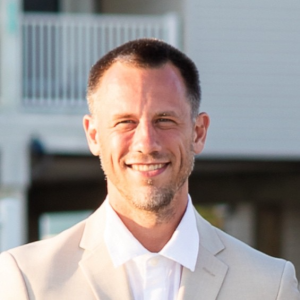 A man with short brown hair and a trimmed beard, wearing a light beige suit jacket over a white shirt, smiles outdoors with sunlight highlighting his face and a blurred building and balcony in the background. Sourcing, Procurement, 3rd Party Risk