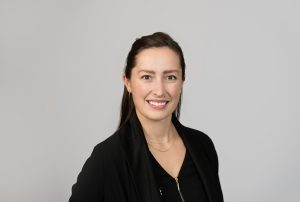 A woman with long brown hair, wearing a black blazer and a black top, smiles confidently at the camera against a simple light gray background. Sourcing, Procurement, 3rd Party Risk