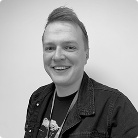 A person with short, styled hair smiles at the camera. They are wearing a dark jacket over a graphic t-shirt and a lanyard. The light-colored, plain background highlights their friendly expression. Sourcing, Procurement, 3rd Party Risk