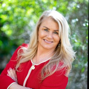 A cheerful woman with long blonde hair, wearing a red jacket, smiles with arms crossed outdoors amid greenery and golden sunlight. Sourcing, Procurement, 3rd Party Risk