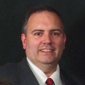 A man in a dark suit, white shirt, and red tie smiles confidently against a black background. Sourcing, Procurement, 3rd Party Risk