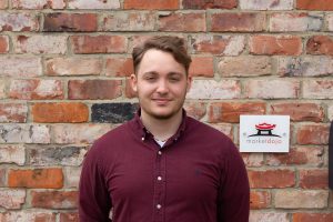 A man with short hair and a trimmed beard wearing a maroon button-up shirt stands in front of a brick wall with a marketdojo sign, highlighting the company’s branding. Sourcing, Procurement, 3rd Party Risk