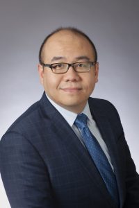 A man with glasses wearing a dark blue checked suit, light blue dress shirt, and blue polka dot tie smiles slightly against a plain gray background, exuding polished professionalism. Sourcing, Procurement, 3rd Party Risk