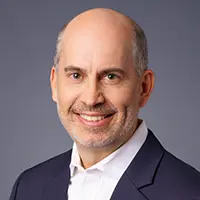 A middle-aged man with a bald head and light facial hair smiles, dressed in a dark suit jacket over a white collared shirt, posed confidently against a plain gray background. Sourcing, Procurement, 3rd Party Risk