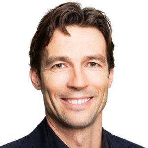 A man with light skin, brown hair, and blue eyes is smiling. He is wearing a dark suit jacket and a dark shirt against a plain white background, creating a professional appearance. Sourcing, Procurement, 3rd Party Risk