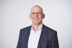 A smiling bald man wearing glasses and a checked suit jacket pairs his look with a light pink plaid dress shirt, standing against a plain light gray background. Sourcing, Procurement, 3rd Party Risk