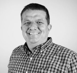 A man with short hair smiles at the camera, wearing a plaid button-up shirt. Photographed against a plain, light-colored background, the black and white image gives a timeless feel. Sourcing, Procurement, 3rd Party Risk