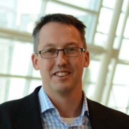 A man wearing glasses, a dark blazer, and a blue checkered shirt smiles in front of large windows with a contemporary, bright background. Sourcing, Procurement, 3rd Party Risk