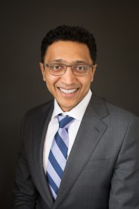 A man with short curly black hair and glasses is smiling. He wears a gray suit, white shirt, and blue striped tie, posing confidently against a plain dark background. Sourcing, Procurement, 3rd Party Risk