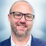 A smiling man with glasses, a beard, and short hair, wearing a white shirt and a blue blazer, poses confidently against a blurred light blue background. Sourcing, Procurement, 3rd Party Risk